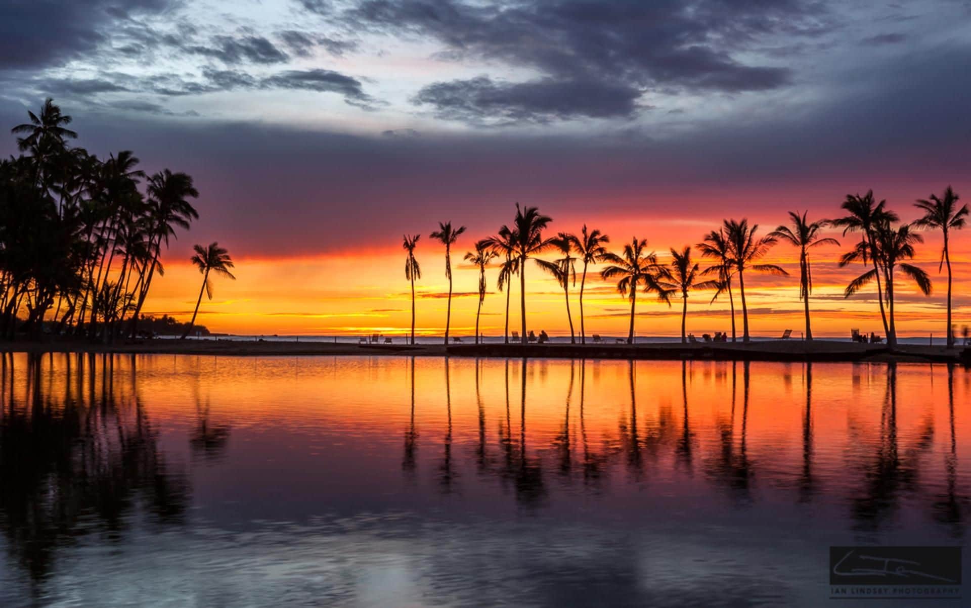 A sunset over a calm beach with palm trees reflected in the water.