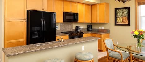 Una cocina con armarios de madera, un refrigerador negro, un microondas, una estufa y un comedor con sillas de mimbre y una mesa de vidrio.