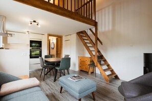 A modern living room with a wooden staircase, a dining area with a table and chairs, and a kitchen with a built-in oven.