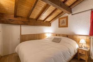 Quarto com teto de madeira, cama com roupa de cama branca, mesa de cabeceira com abajur e quadro na parede.