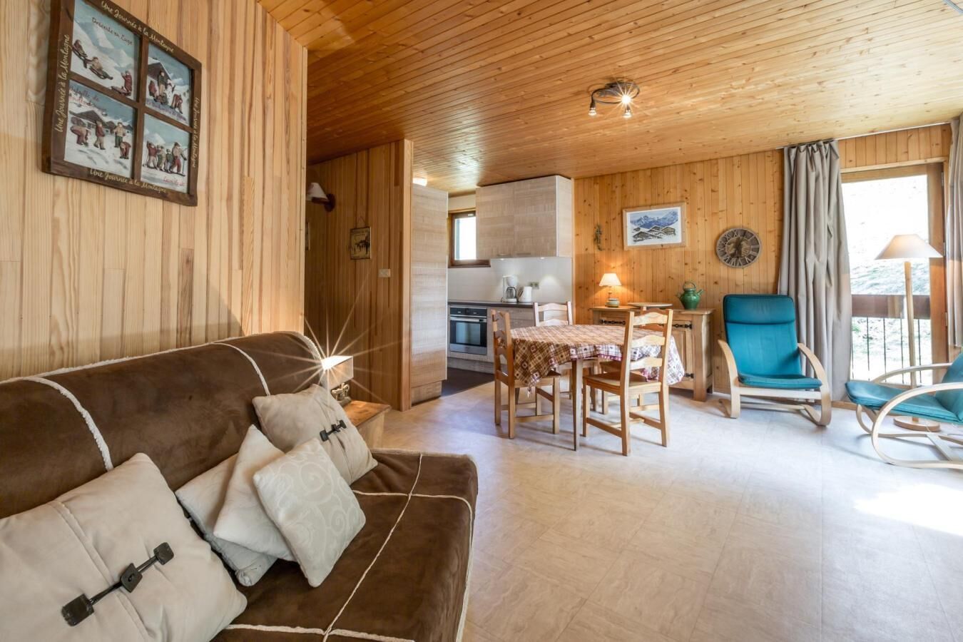 Una habitació acollidora amb un sostre de fusta, un sofà marró amb coixins, una taula de dinar amb cadiretes i una cuina al fons.