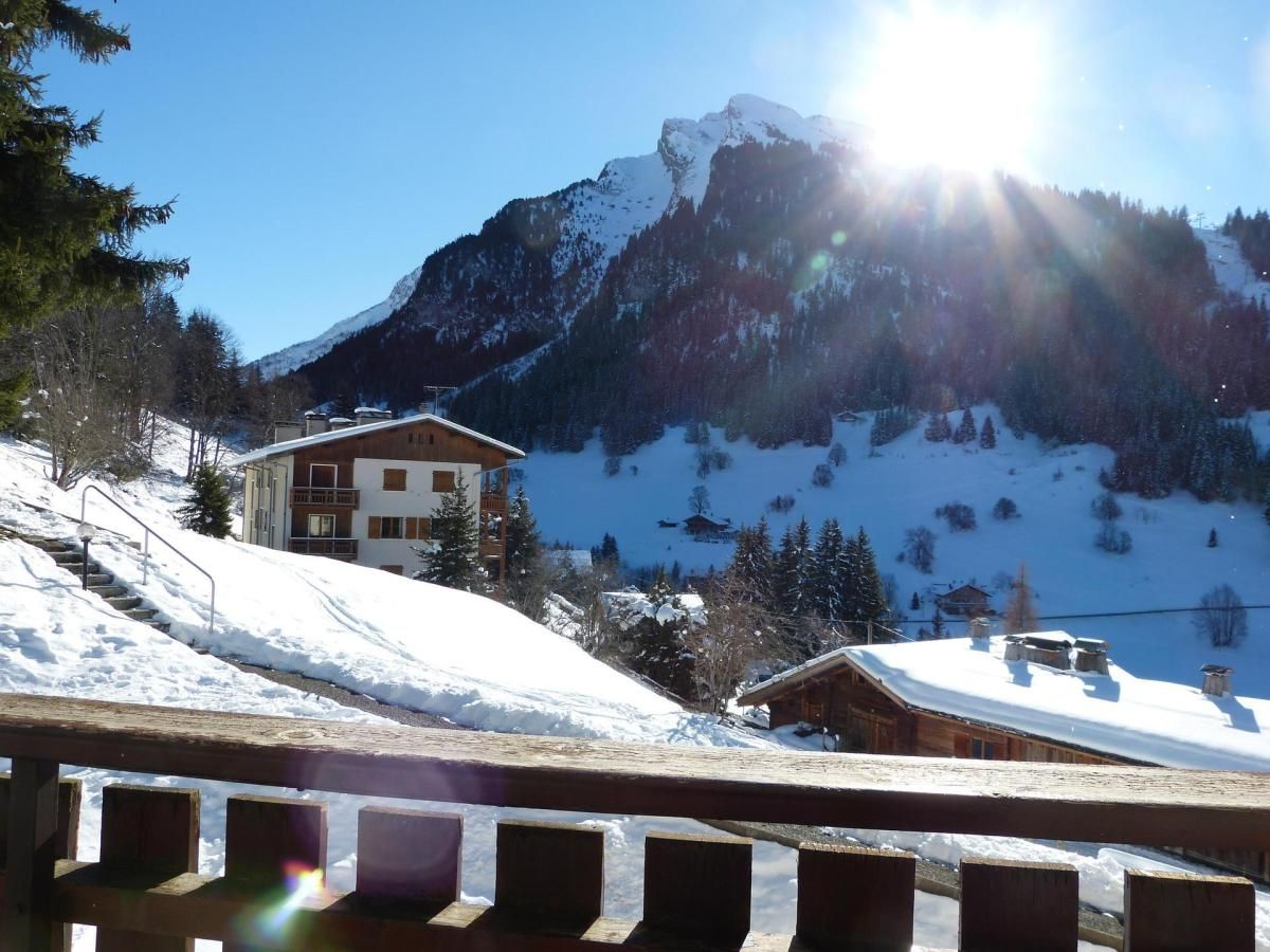 A wooden chalet with a snow-covered roof, a ski slope in the background, and a mountain range.
