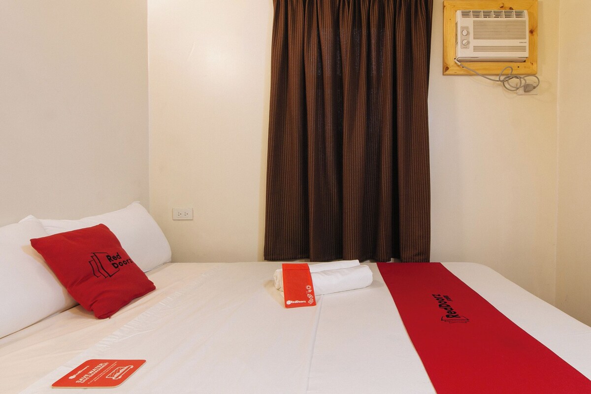 Ein Hotelzimmer mit einem Bett, einer rot-weißen Tagesdecke, einem roten Kopfkissen, einem weißen Kopfkissen, einer rot-weißen Decke, einer Wand-Klimaanlage und einem Fenster mit braunen Vorhängen.