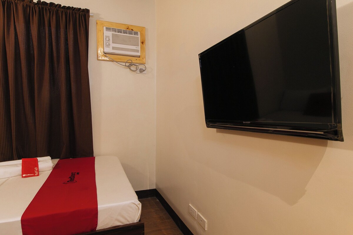 Un lit simple avec une couvre-lit rouge et blanche, un téléviseur fixé au mur, une fenêtre avec des rideaux et un climatiseur mural.