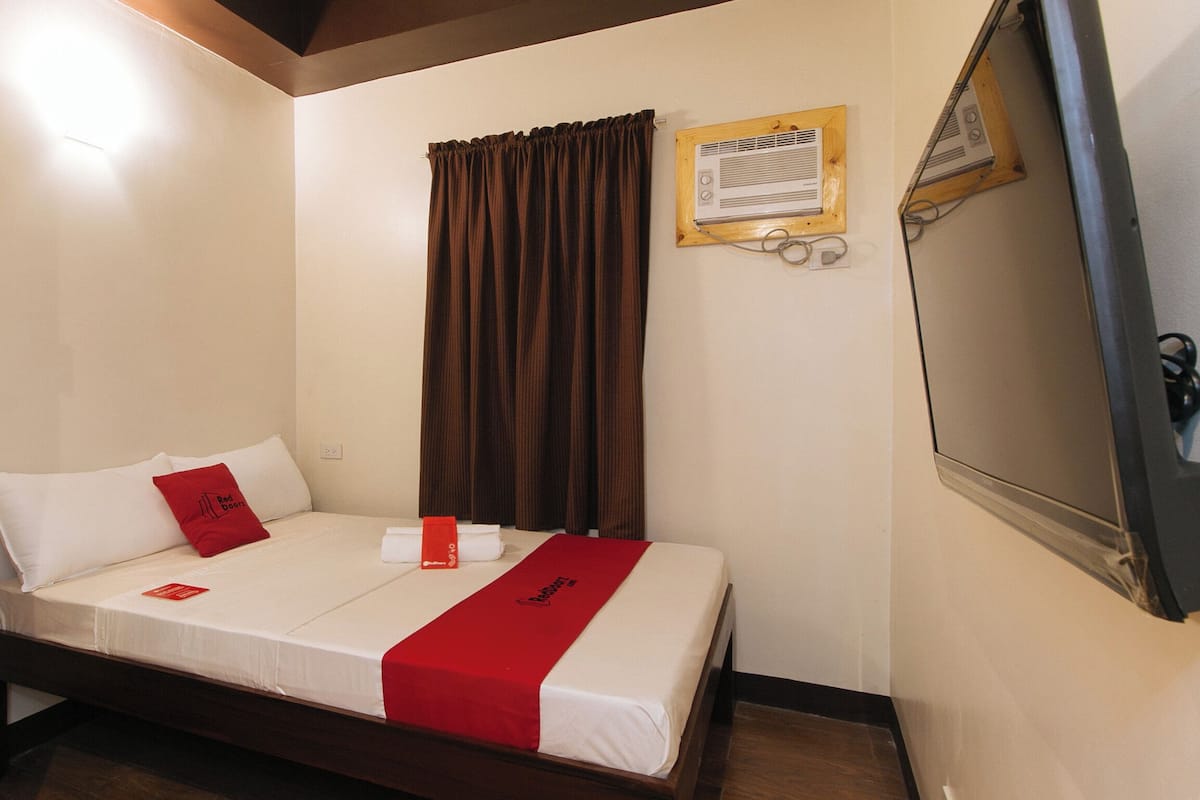 Habitación de hotel con cama, mesita de noche, espejo, ventana con cortinas y aire acondicionado instalado en la pared.