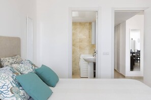 Quarto com cama, almofadas, banheiro com pia e área de jantar visível por uma porta aberta.