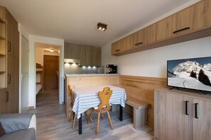 A compact kitchen with wooden cabinets, a dining table with chairs, a bench, and a television on a wooden cabinet.