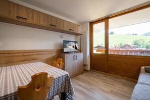 Ein Zimmer mit einem Holztisch, Stühlen, einem Fernseher und einem großen Fenster mit Blick auf eine grüne Landschaft.