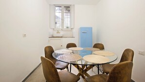 A compact dining area with a round glass table and brown upholstered chairs. A blue refrigerator is visible in the background, and a window with a view of greenery is above the sink. - St. Agnes Hannibal (Roma)