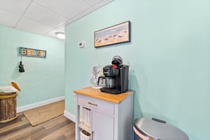 A kitchenette with a coffee maker, a trash bin, a wicker chair, and a wall-mounted picture. - 2BR Condo Sleeps 6 • Pool & Steps to Beach (Panama City Beach)