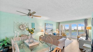 A spacious living room with a glass dining table, wicker chairs, a sofa, and a ceiling fan. - 2BR Condo Sleeps 6 • Pool & Steps to Beach (Panama City Beach)