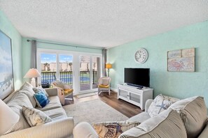 A living room with a sofa set, a television, a clock, and a view through a glass door.