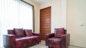A room with two red leather sofas, a wooden door, and a large window with blinds. - RedDoorz near ITDC Nusa Dua (Bali)