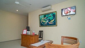 A reception area with a wooden desk, a chair, a table with a lace tablecloth, a painting, and a television mounted on the wall. - RedDoorz near ITDC Nusa Dua (Bali)