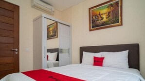 A hotel room with a bed, a red throw, a painting, and a wooden door. - RedDoorz near ITDC Nusa Dua (Bali)