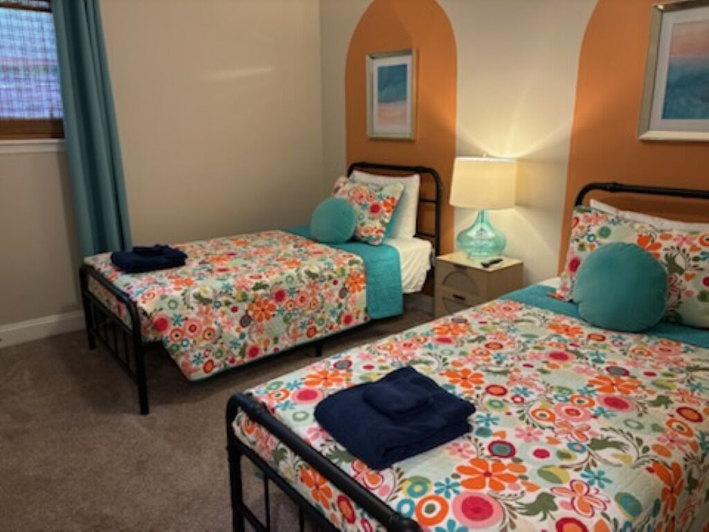 Een kamer met twee eenpersoonsbedden, elk voorzien van bloemenbeddels en teal kussens. Daartussen bevindt zich een nachtkastje met een lamp.