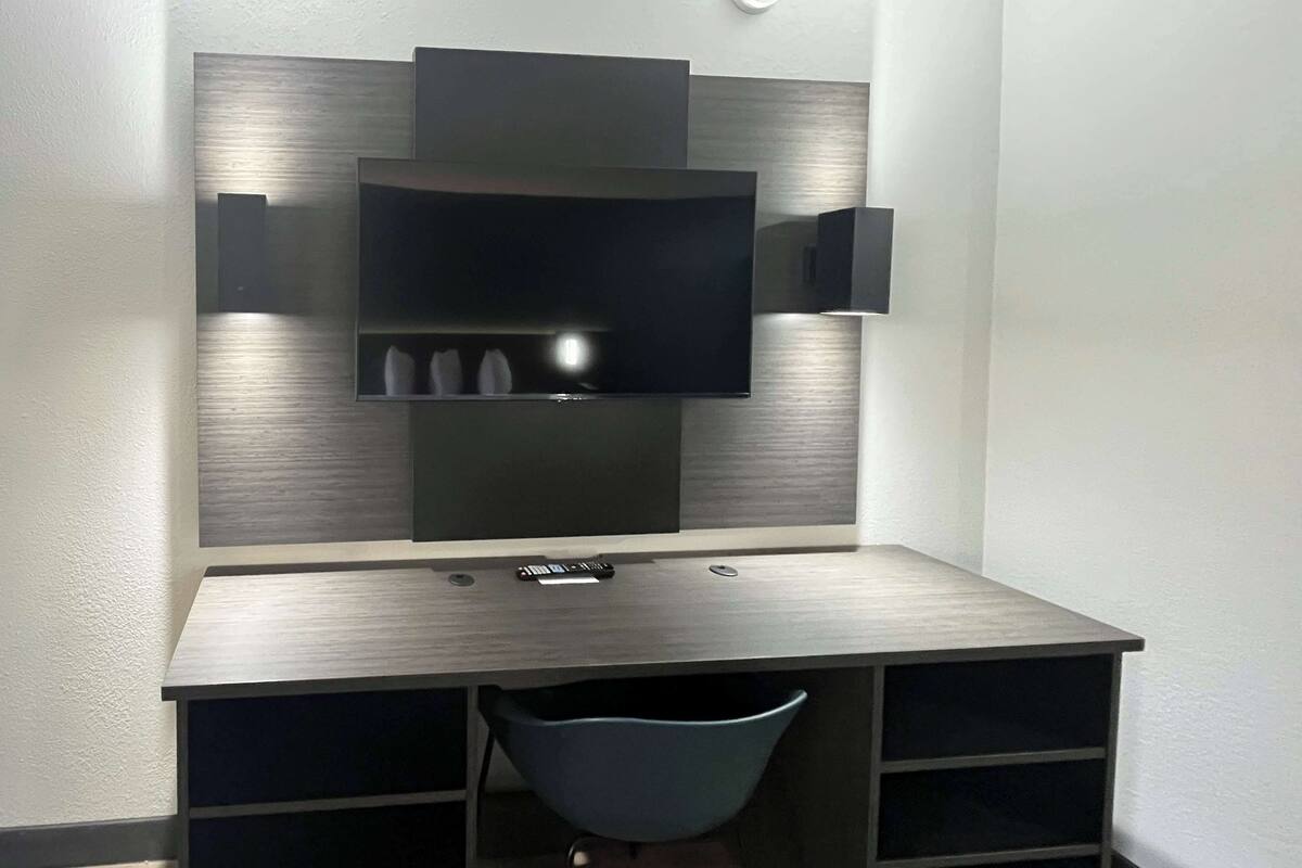 A modern, dark-colored desk with a flat-screen TV mounted on the wall above it. There are two remote controls on the desk, and a blue chair is positioned in front of the desk.