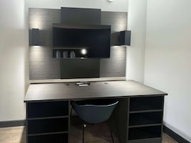 A modern, dark-colored desk with a flat-screen TV mounted on the wall above it. There are two remote controls on the desk, and a blue chair is positioned in front of the desk.
