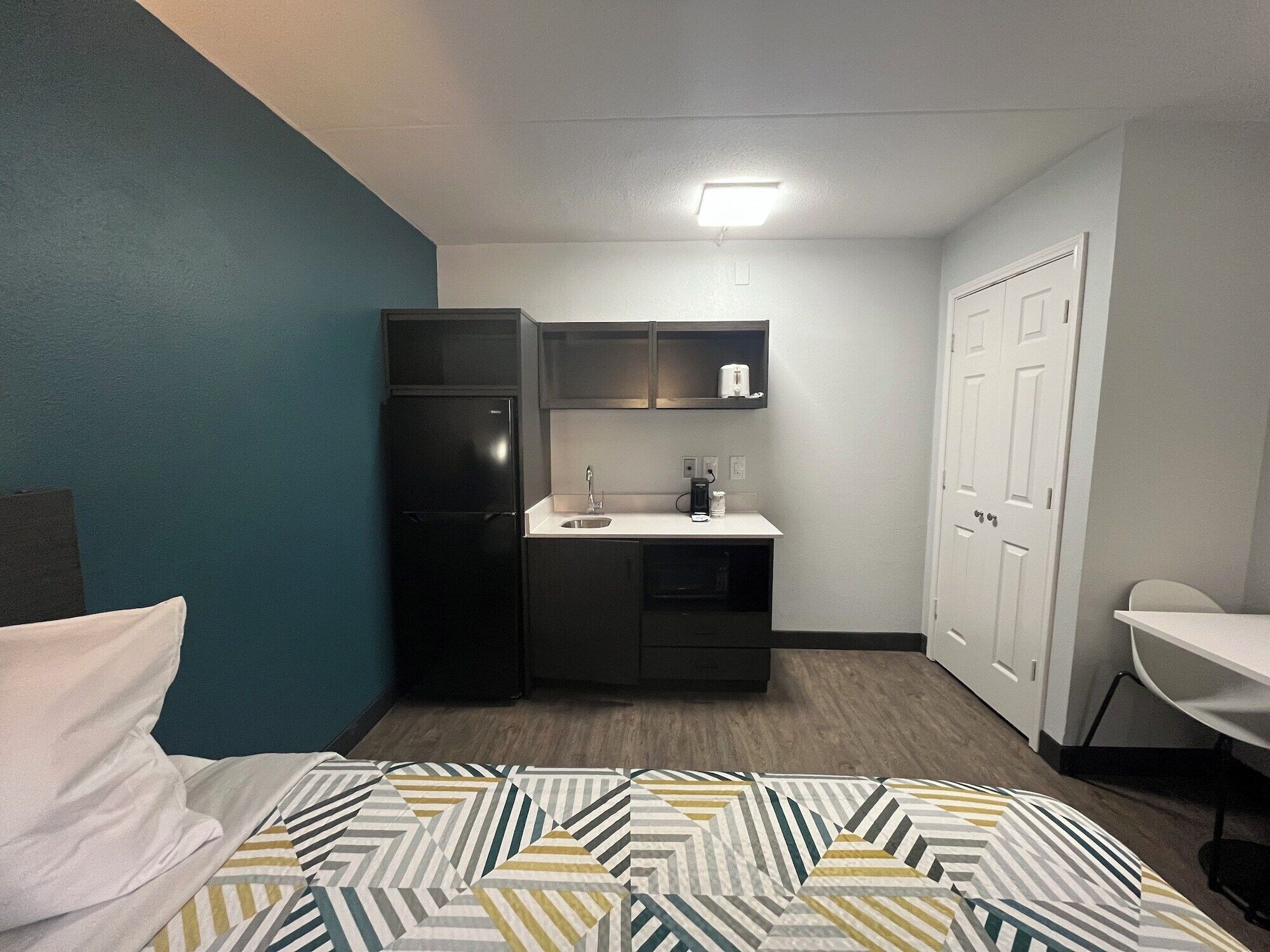 A compact hotel room with a bed, a kitchenette, a desk, and a chair.