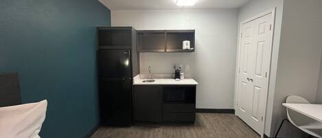 A compact hotel room with a bed, a kitchenette, a desk, and a chair.