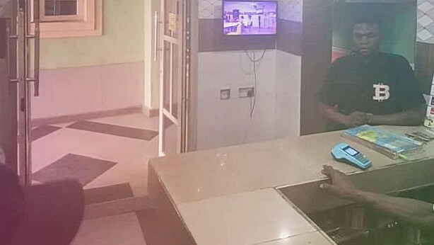 Zona de recepción con un reloj, un televisor y un mostrador donde hay una persona sentada.