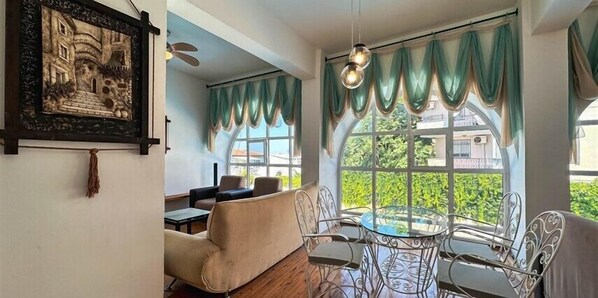 Una habitación con comedor que incluye una mesa redonda de vidrio y sillas de metal, un sofá y un ventanal con cortinas verdes.