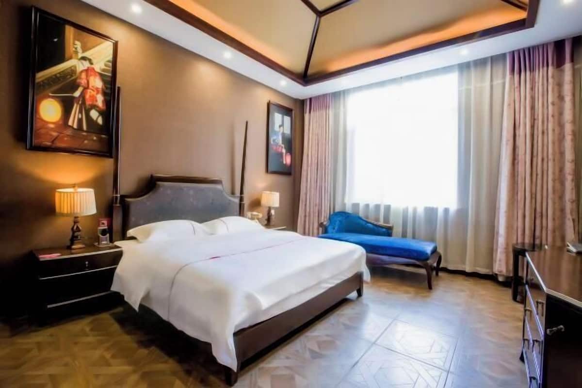 Une chambre d’hôtel avec un lit, des tables de chevet, une commode, un canapé bleu, une fenêtre avec des rideaux et des œuvres d’art accrochées aux murs.
