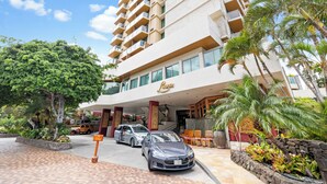 Un hôtel au design moderne, avec une entrée imposante, un parking où sont garées des voitures et un aménagement paysager luxuriant de style tropical.