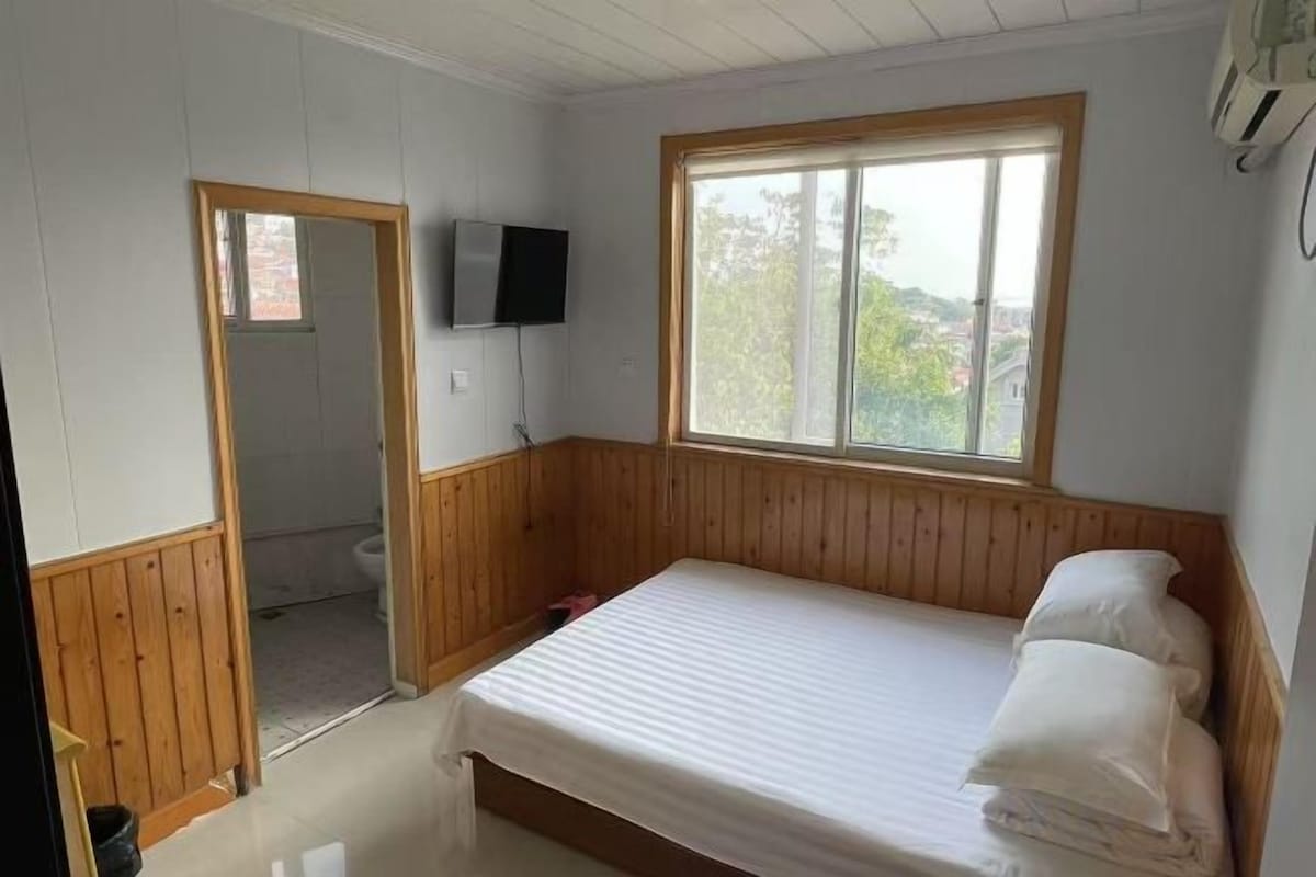 Habitación individual con una cama y cabecera de madera, ventana con vistas, televisor montado en la pared y puerta que da al baño.
