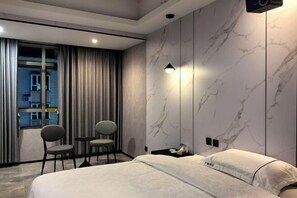 A modern hotel room with a large bed, two chairs, a small table, and a view of the outside through a large window.