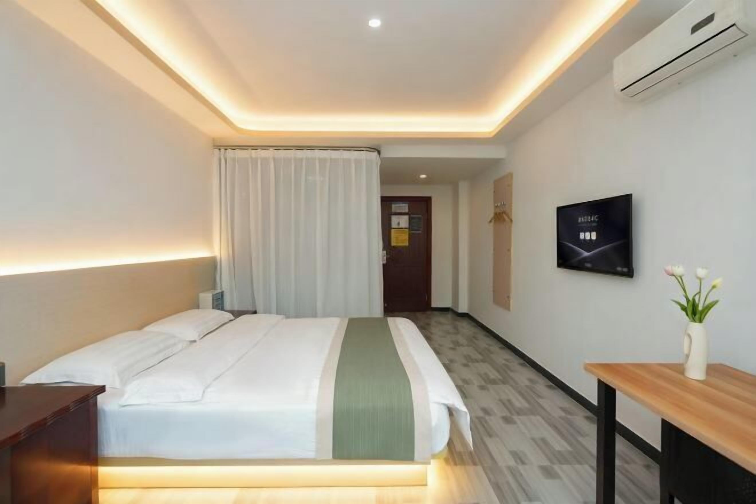 A hotel room with a bed, a bedside table, a TV mounted on the wall, and a wooden bench.