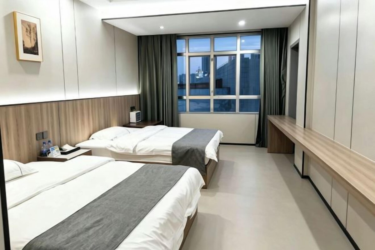 A hotel room with two beds, a bedside table, a nightstand, a window with curtains, and a view of the city.