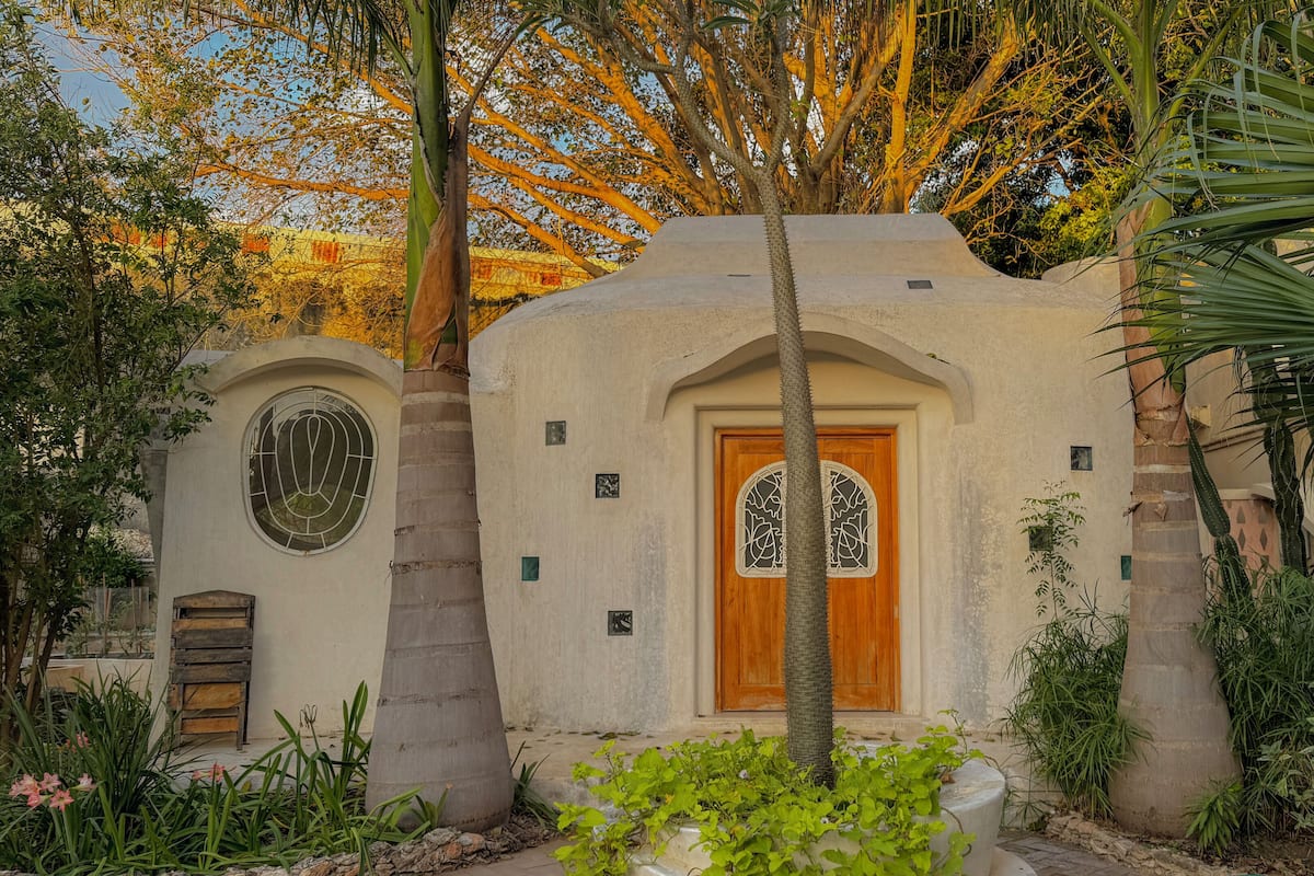 Un edificio con puerta de madera y ventana circular, rodeado de plantas y árboles.