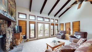 A spacious living room with a stone fireplace, large windows, and a ceiling fan. - Bugle Point - Unit I1 (Ruidoso)