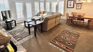 Un salon comprenant un canapé, une table basse et une table à manger. De grandes fenêtres sont équipées de stores, et on y trouve également un tapis et un lustre.