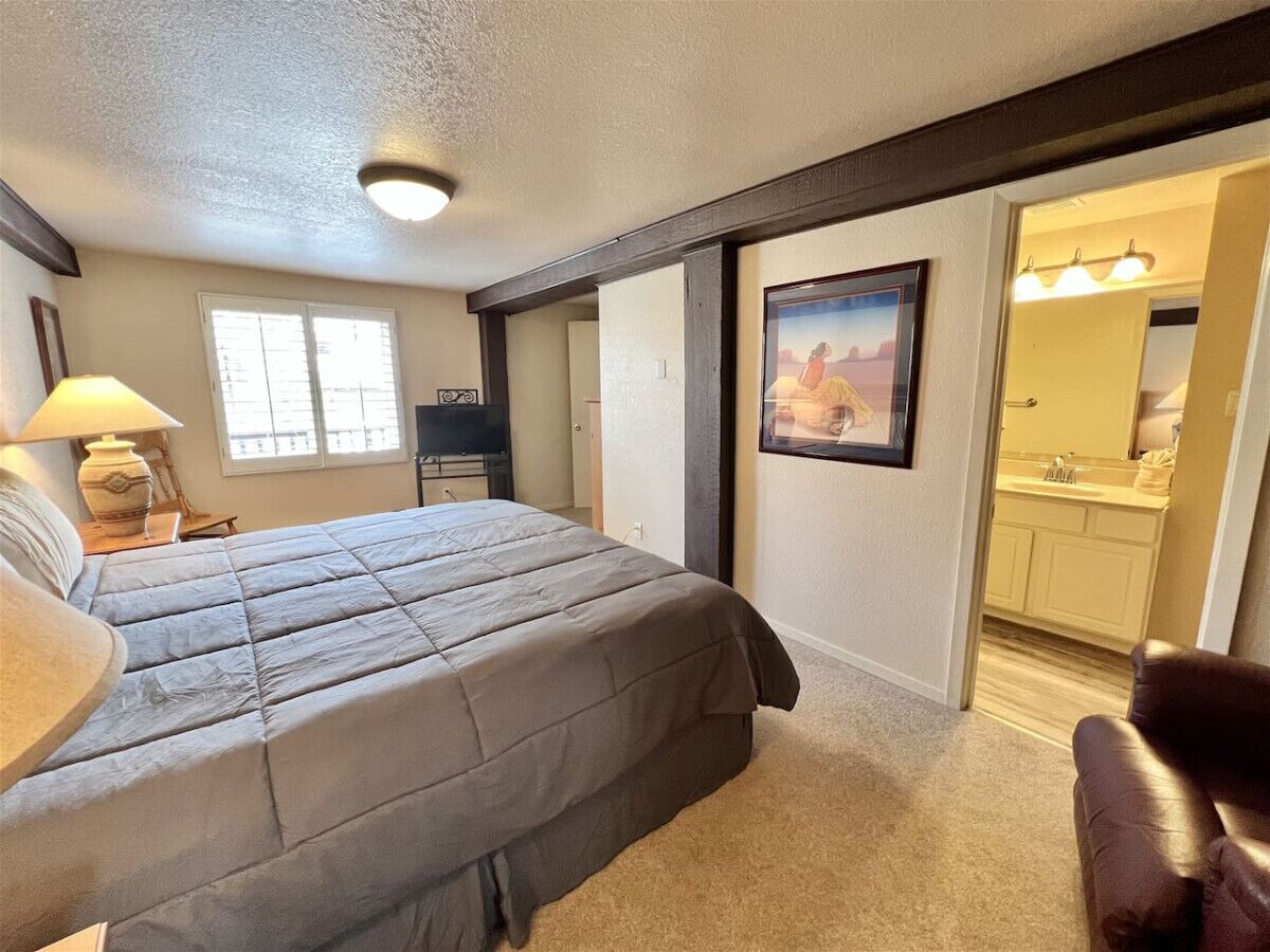 A bedroom with a large bed, a desk with a lamp, a chair, a painting on the wall, and a bathroom visible through an open door.
