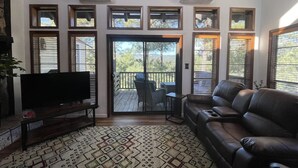 A living room with a large flat-screen TV, a leather sofa, a patterned rug, and a balcony with a view of trees.