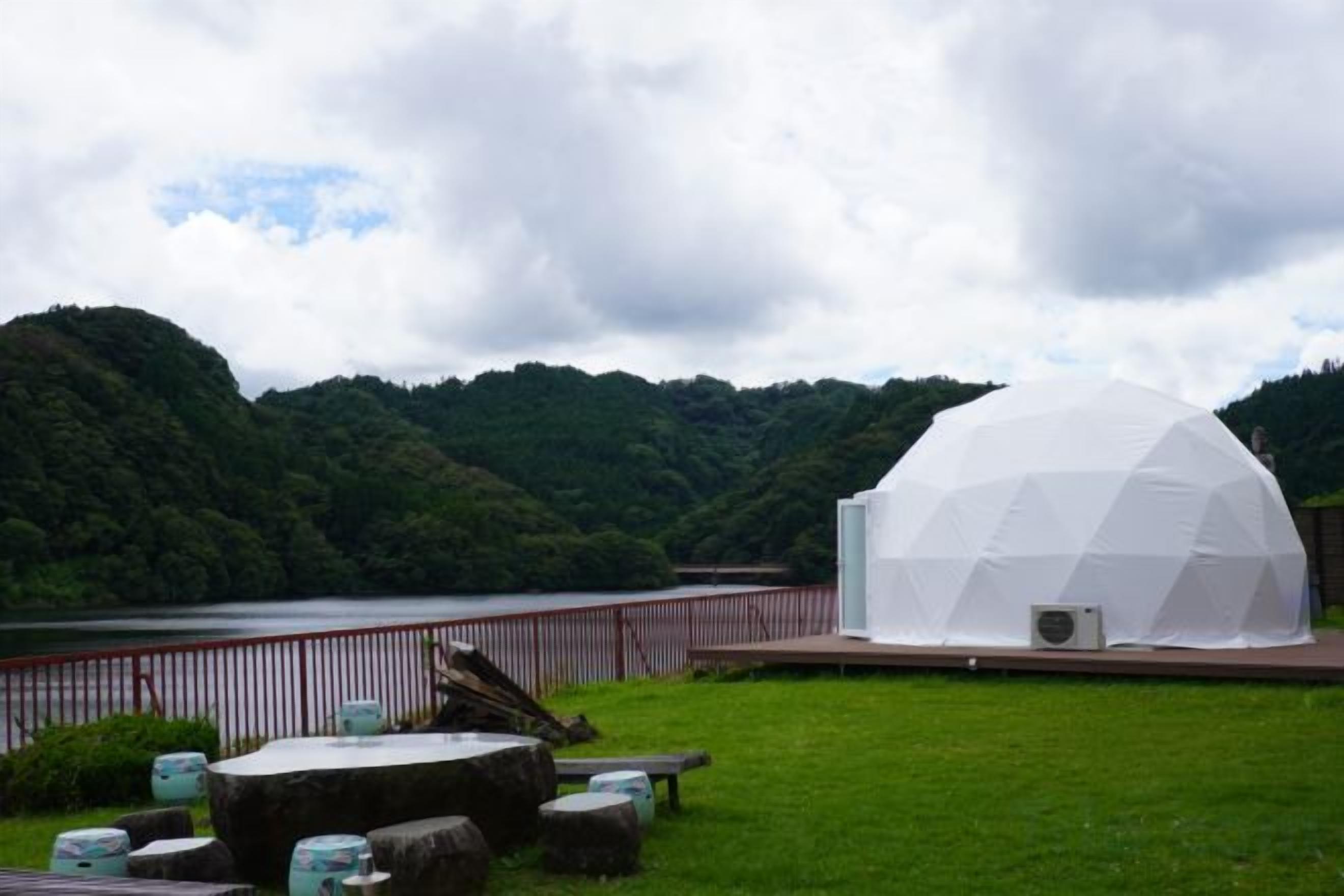 A dome-shaped structure with a flat roof, surrounded by a grassy area with a lake and forested hills in the background.