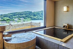 Un onsen japonés tradicional con una bañera de madera y un baño de aguas termales, con vistas panorámicas a un río y colinas.