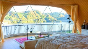 Ein Schlafzimmer mit Bett, kleinem Tisch und Stuhl, einem Fenster mit Seeblick und Hügeln sowie einem Balkon mit Geländer.
