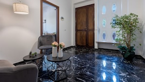 Un interior moderno con suelo de mármol, un sillón gris, una mesa redonda con jarrones y una puerta de madera con ventanas hexagonales.