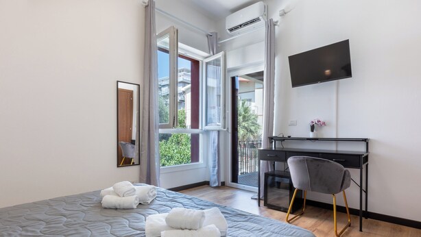 Habitación de hotel con una cama grande, un escritorio, una silla, un televisor y un balcón con vistas a edificios y árboles.