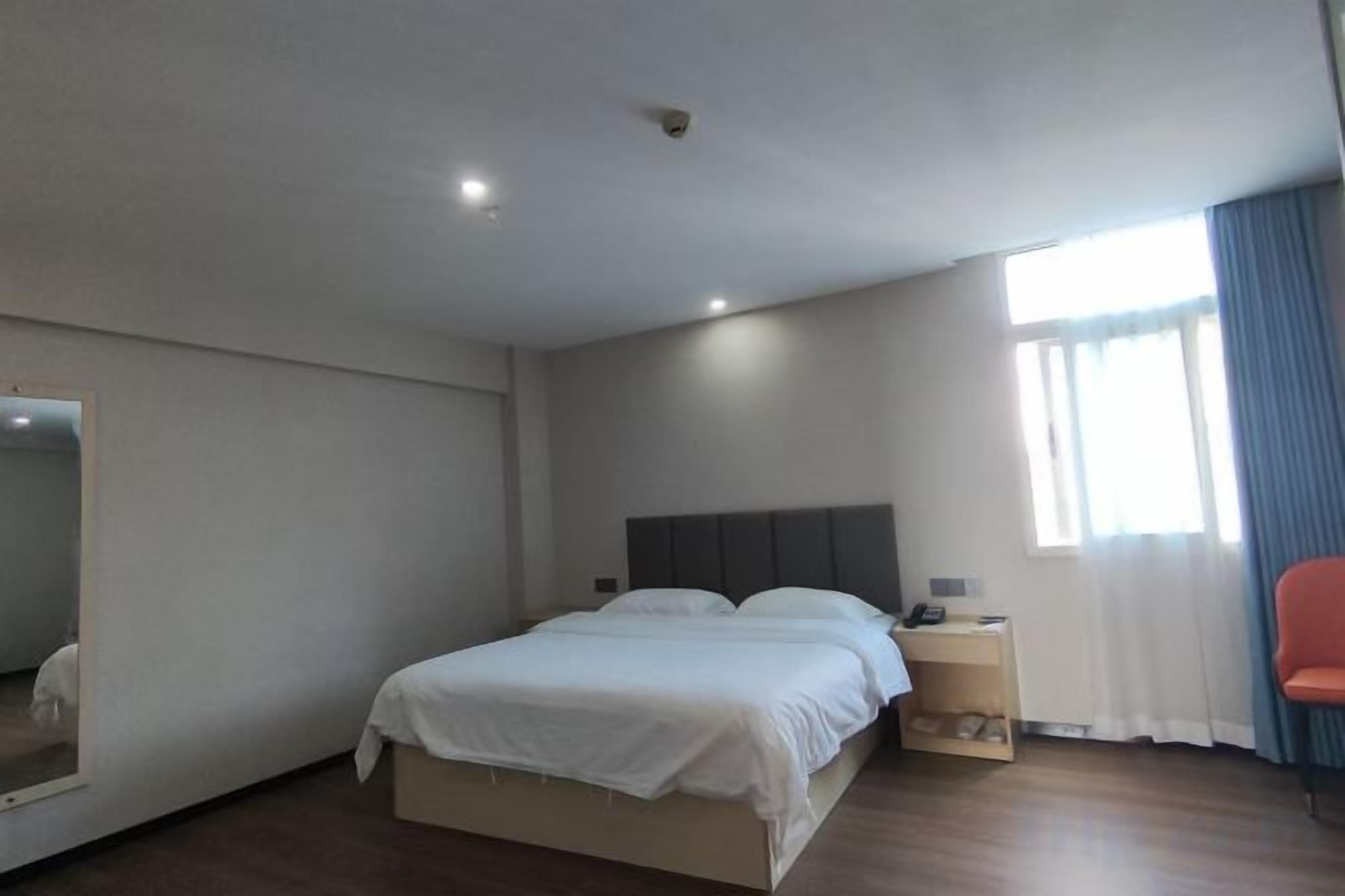 A hotel room with a bed, a bedside table, a chair, a window with curtains, and a mirror on the wall.