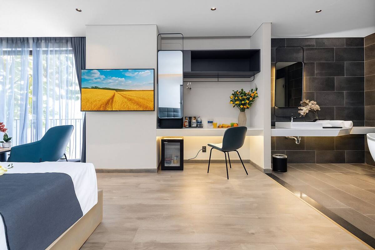 Une chambre d’hôtel moderne équipée d’un lit, d’un bureau, d’une chaise, d’une télévision, d’un minibar, d’une salle de bain avec un lavabo et un miroir, et d’une grande fenêtre avec des rideaux.