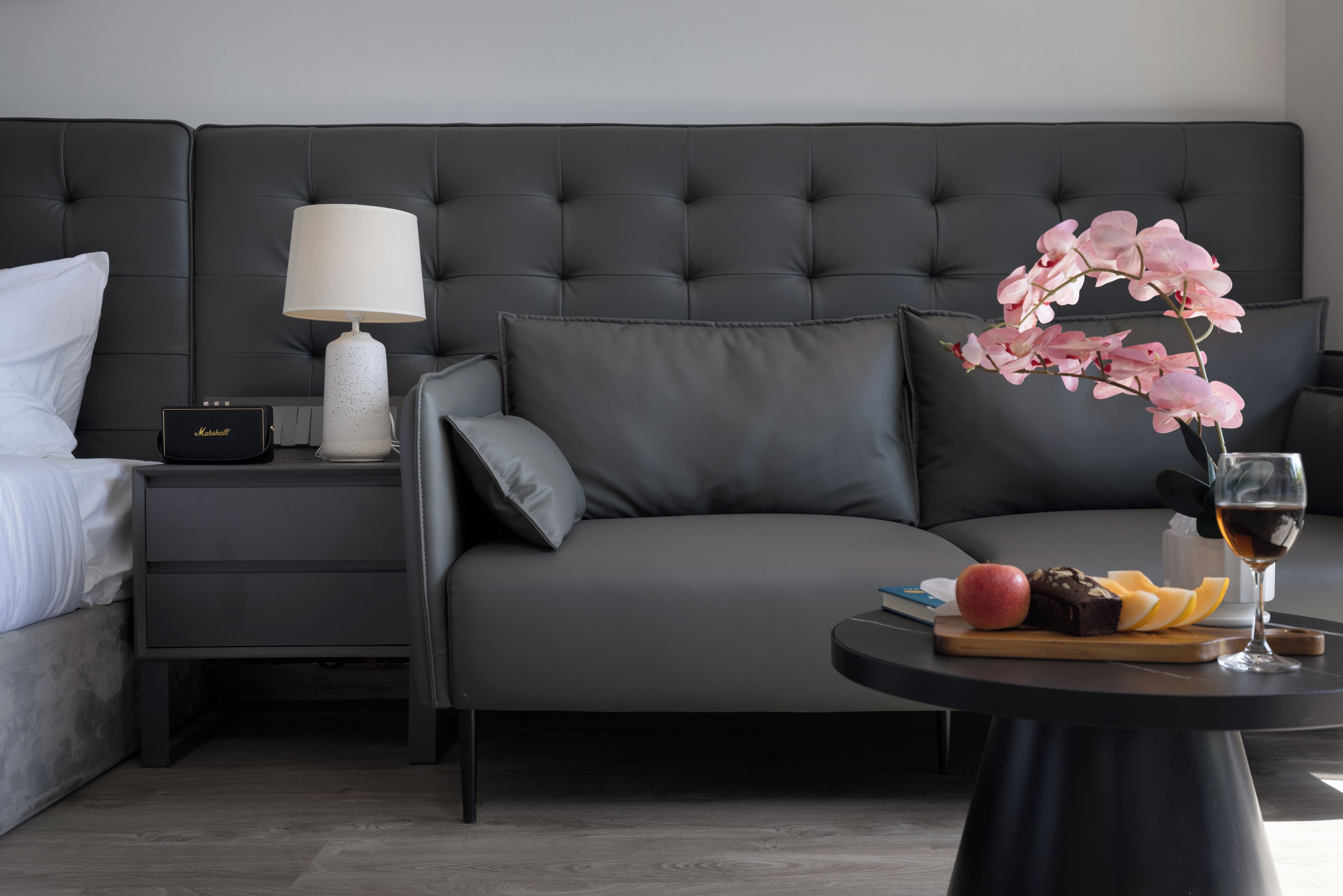 Et moderne hotelværelse med en grå sofa, et lille natbord med en lampe, et sengebord og et rundt bord med frugt og et glas vin.