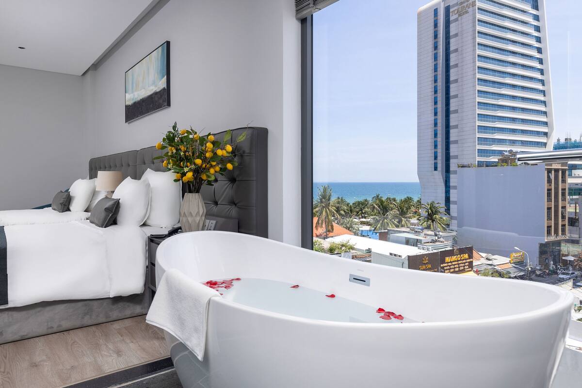 Une chambre d’hôtel moderne dotée d’un grand lit, d’une baignoire remplie de pétales de rose, d’une vue sur l’océan et sur le paysage urbain.