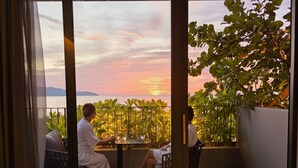 En balkon med to personer siddende, med udsigt over en solnedgang med vand og grønt.