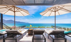A balcony with two lounge chairs and a table, overlooking a clear blue sea and distant mountains. - JBAY Beachfront Boutique Hotel  (Da Nang)