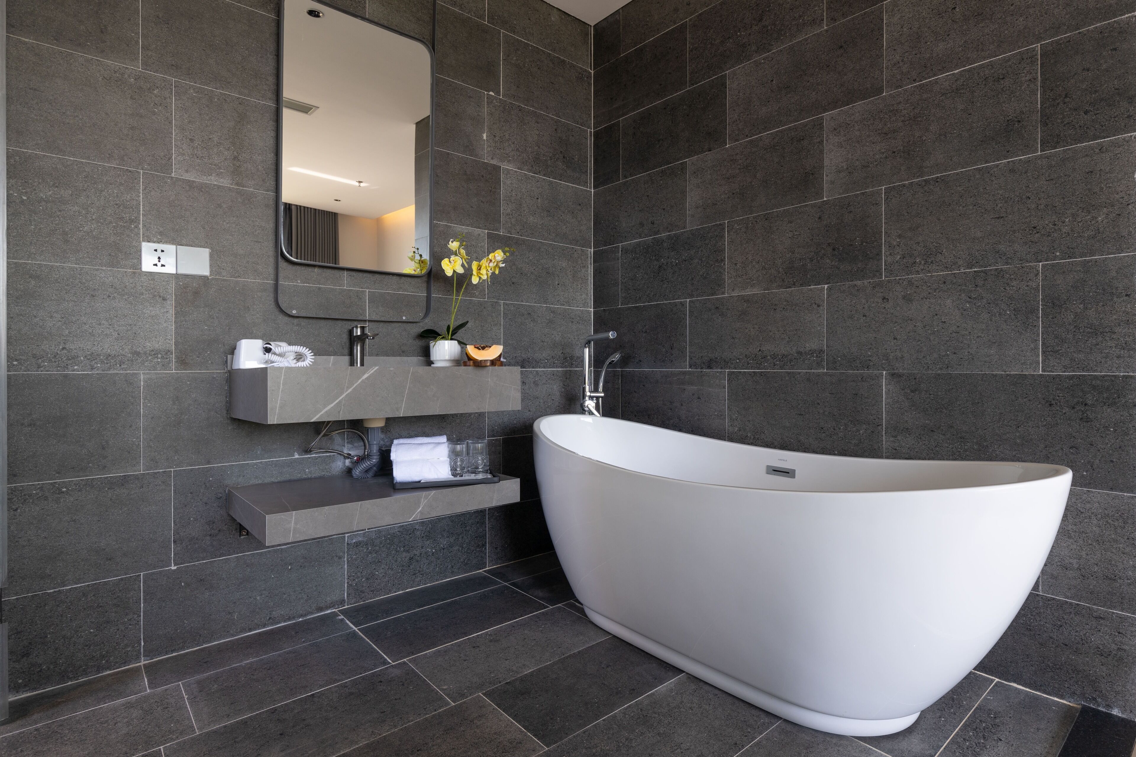 Ein modernes Badezimmer mit dunklen Fliesenwänden, einer freistehenden Badewanne, einem Waschtisch mit Spiegel und einem kleinen Topfpflanzen.