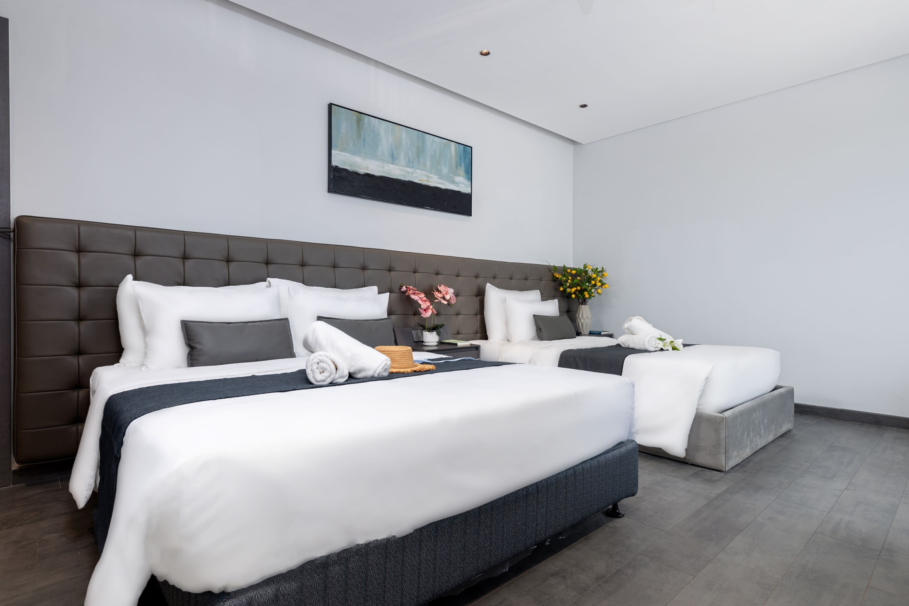 Une chambre d’hôtel moderne avec deux lits, une tête de lit grise, du linge de lit blanc, un tableau au mur et un vase avec des fleurs.